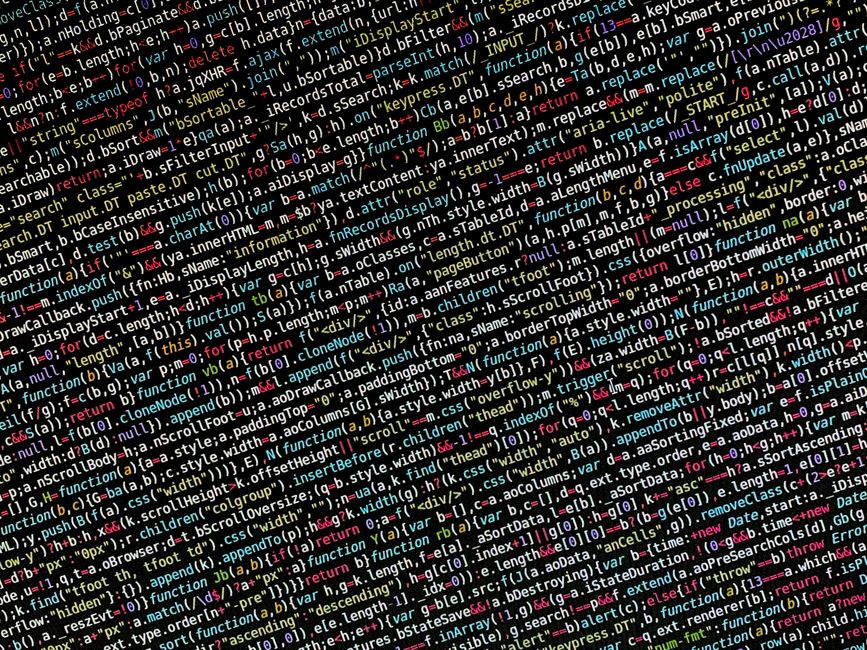 A person working on a laptop with a dark background, displaying lines of computer code in multiple colors, sitting at a black table. The individual's hands are visible, with one hand typing on the key