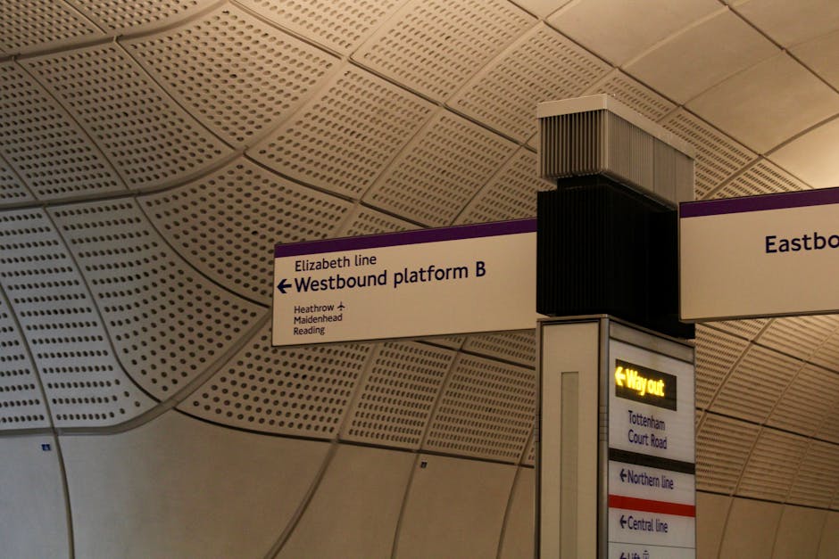 Inside a modern underground train station with a curved, perforated ceiling made of perforated metal panels in a grid pattern, illuminated by soft lighting. Prominent white and purple directional sign